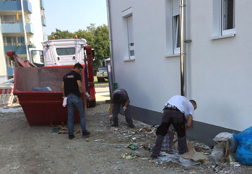 Drei Arbeiter räumen Müll und Bauschutt neben einem Gebäude auf, während ein großer roter Container im Hintergrund steht.