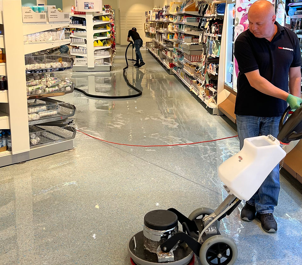 Zwei Reinigungskräfte in einem Supermarkt. Eine Person nutzt eine Bodenreinigungsmaschine, während die andere mit einem Schlauch den Boden säubert.