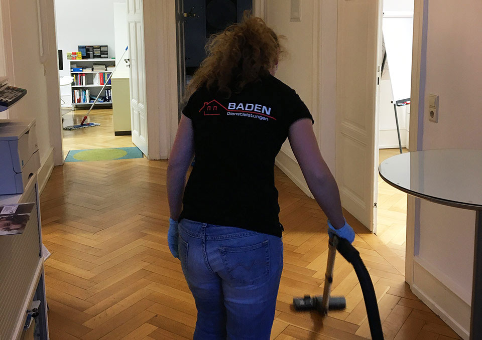 Frau mit lockigem Haar in schwarzem T-Shirt mit Aufdruck "Baden" reinigt den Boden mit einem Staubsauger in einem Büro.
