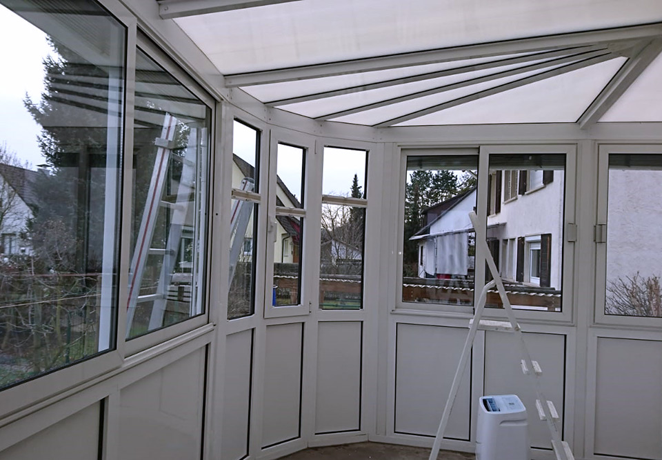 Sauberer Wintergarten mit großen Fenstern, einem weißen Luftentfeuchter und einer Leiter im Hintergrund.