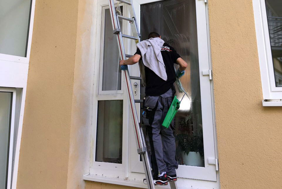 Ein Fensterputzer steht auf einer Leiter und reinigt ein Fenster. Er trägt ein schwarzees T-Shirt und eine graue Hose, mit einem Reinigungstuch über der Schulter.