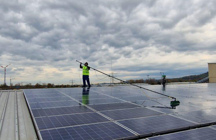 Arbeiter reinigt Solarpanels auf einem Dach mit einem langen Besen, bei bewölktem Himmel.