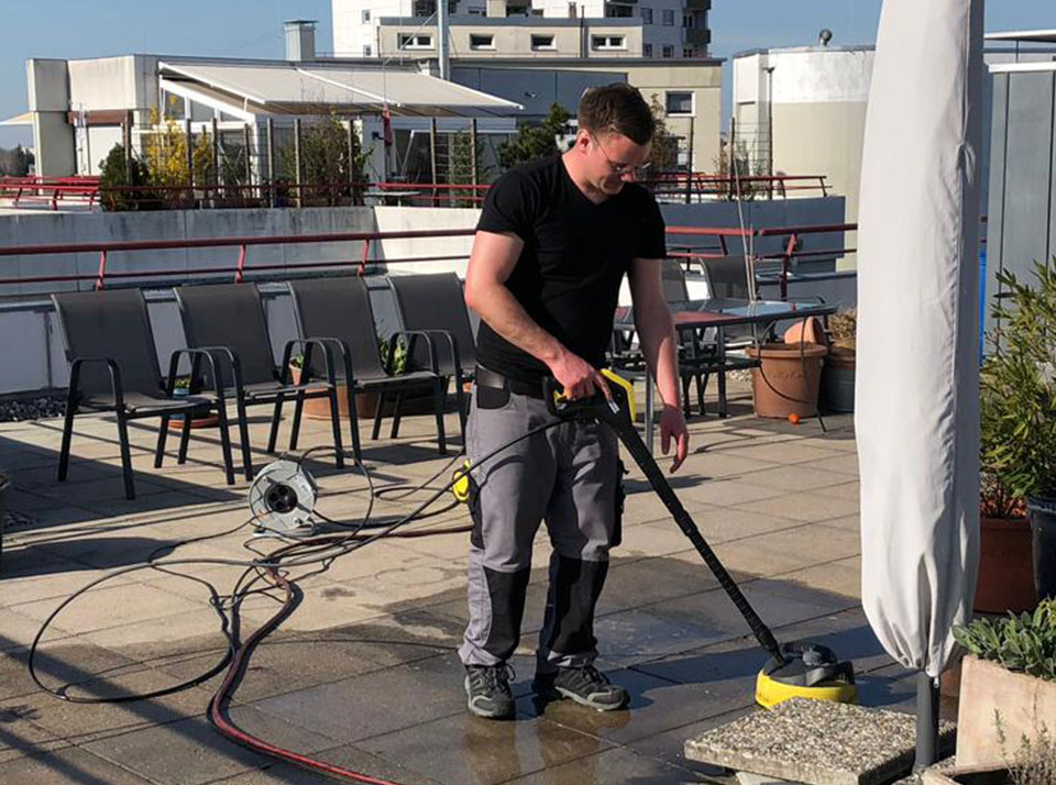 Mann mit Kurzarmshirt und Arbeitshose reinigt mit Hochdruckreiniger eine Terrasse. Im Hintergrund sind Stühle und Pflanzen zu sehen.