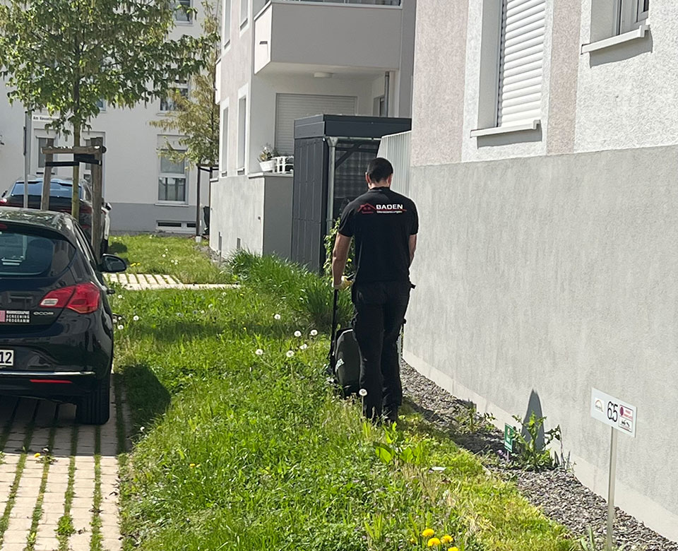 Mann mit Rasenmäher schneidet Gras neben einem Wohngebäude. Im Hintergrund ein schwarzes Auto und eine gepflegte Umgebung.